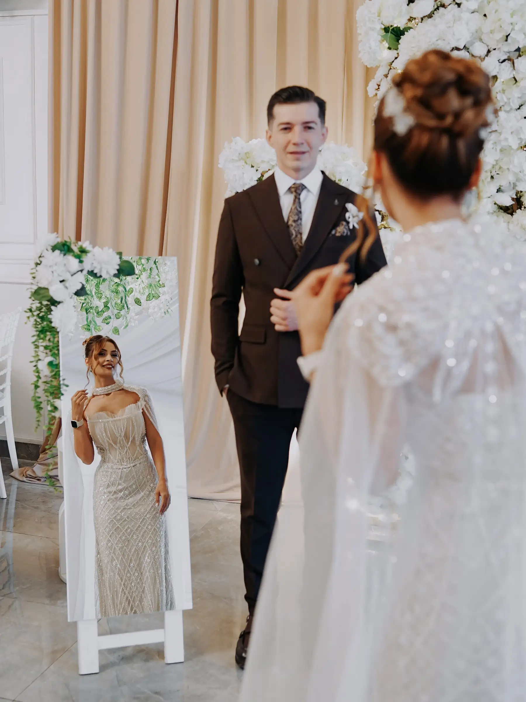 Damat gelini karşılıyor; gelin sırtı dönük, pırıltılı pelerinli elbisesi ve boy aynasındaki yansıması görünüyor