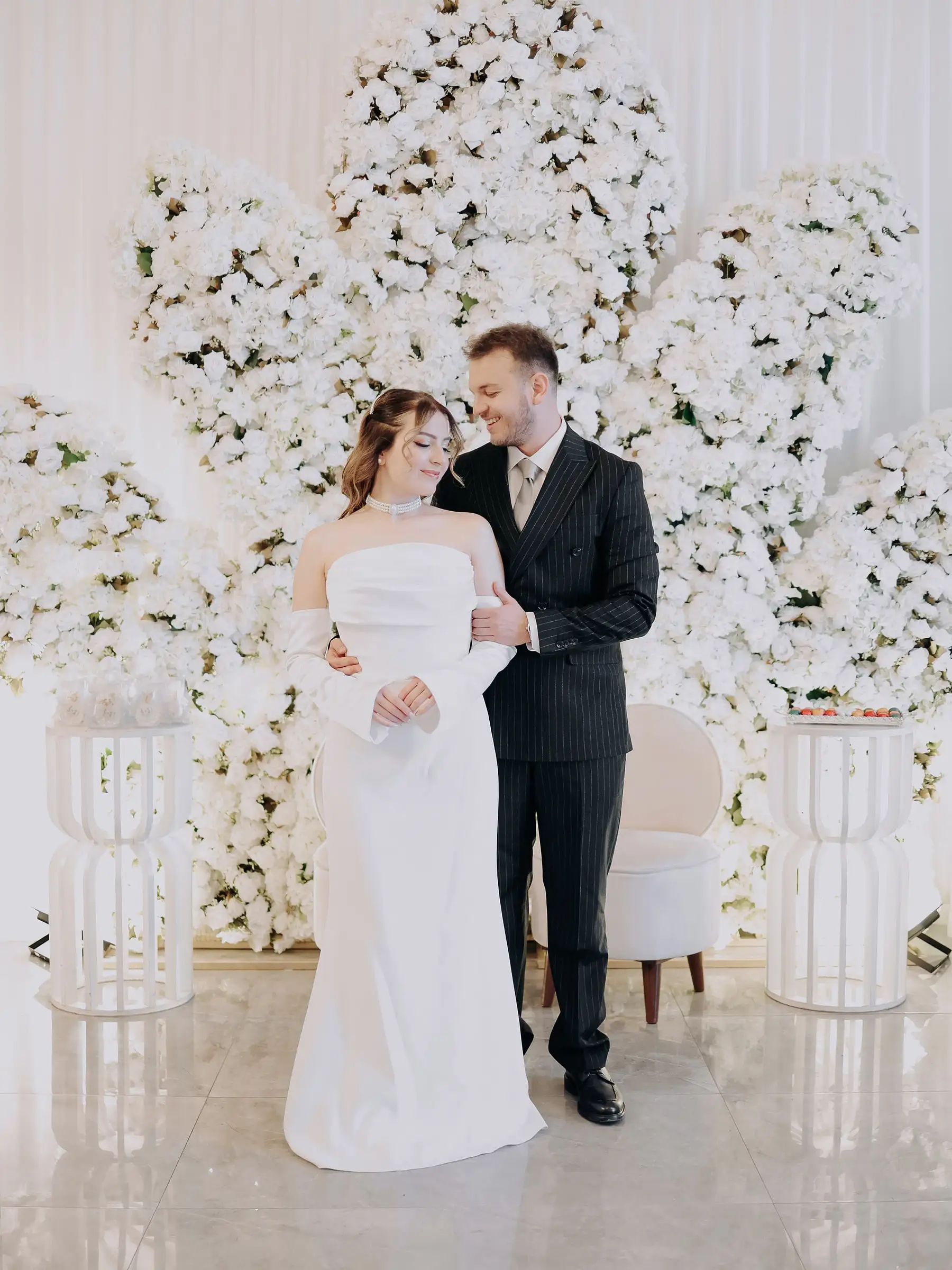 Gelin ve damat, beyaz çiçek duvarının önünde birbirine sarılıyor; gelin sade beyaz elbiseli, damat çizgili takım elbiseli