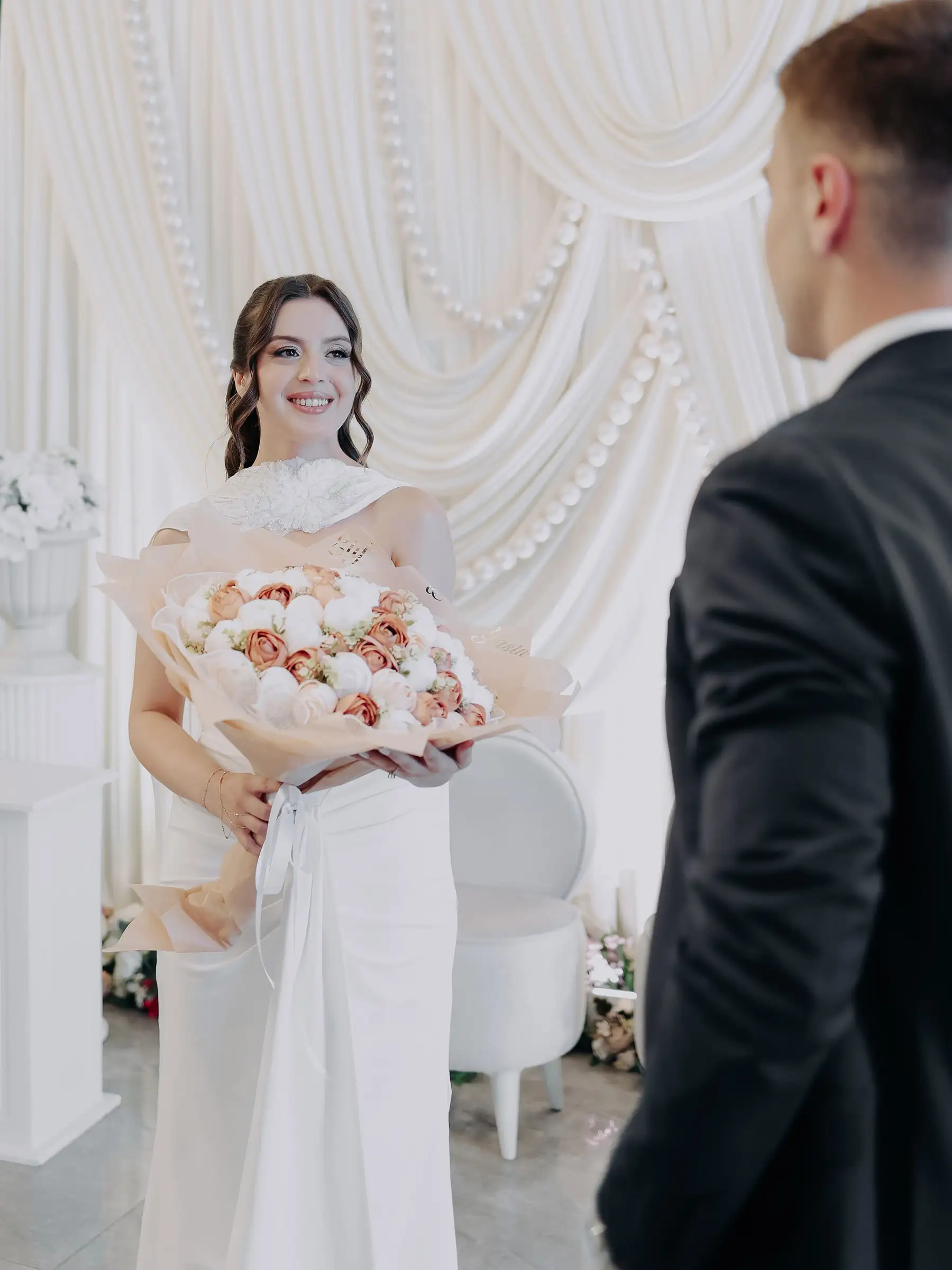 Gelin yumuşak renkli buketini tutuyor, damat sağ kenarda kısmen görünüyor; arkada beyaz drapeli fon