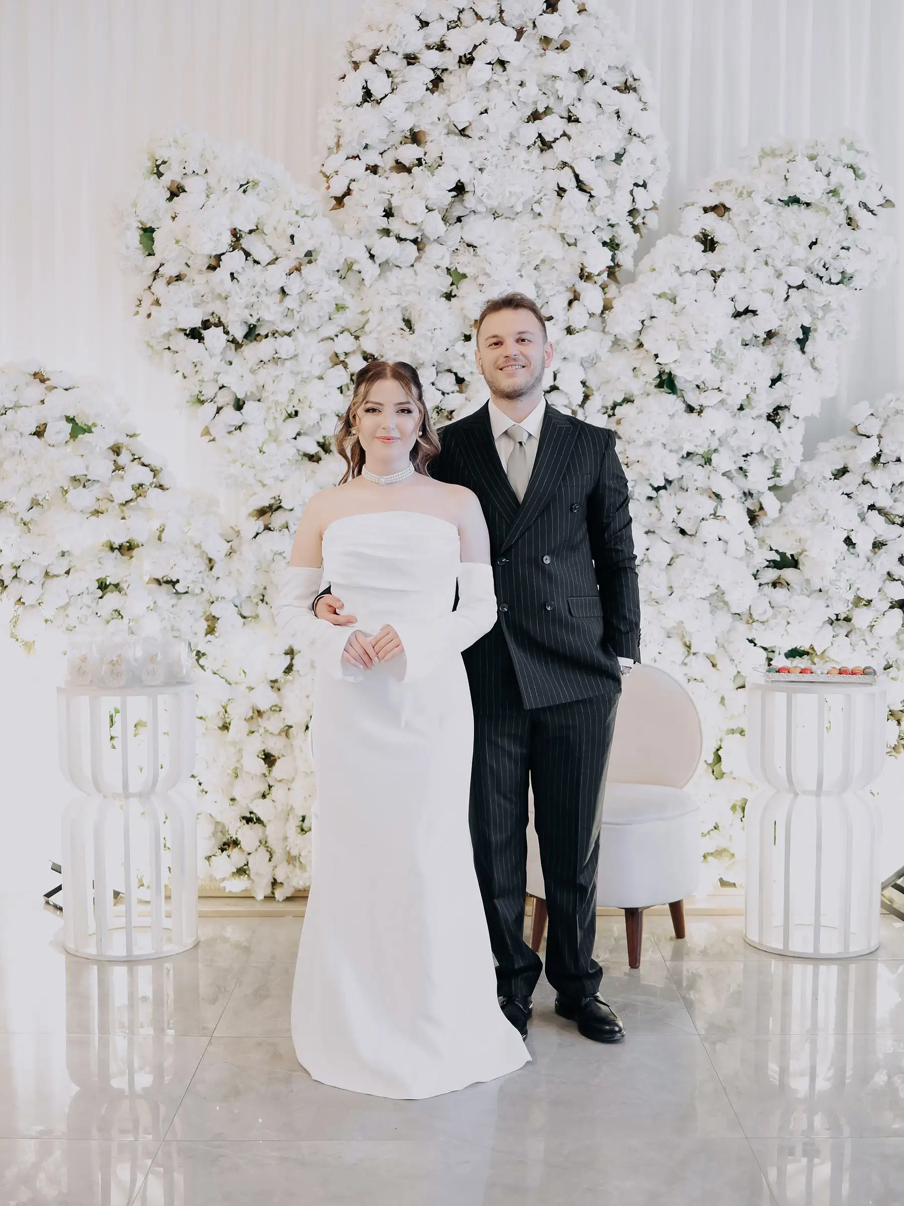 Gelin ve damat, dev beyaz çiçek duvarının önünde yan yana poz veriyor