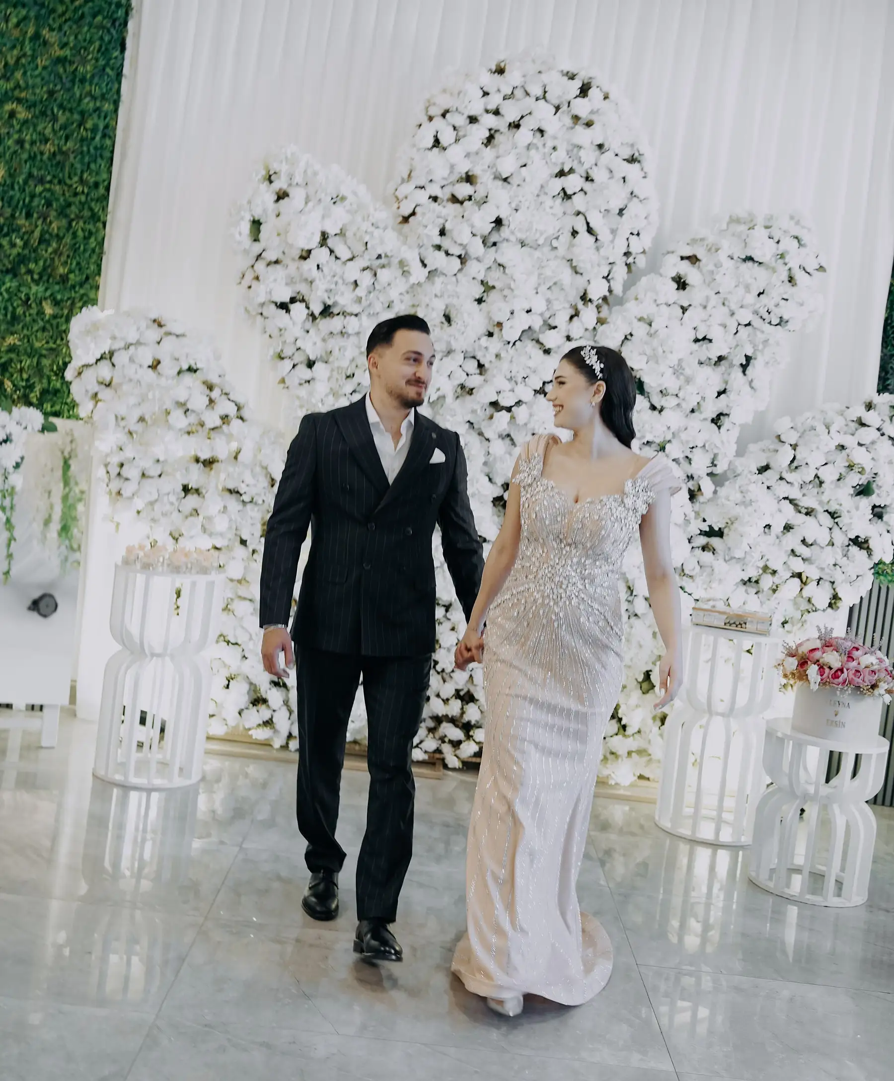 Gelin ve damat el ele yürüyor; arkada beyaz çiçek duvarı ve çiçekli sehpalar