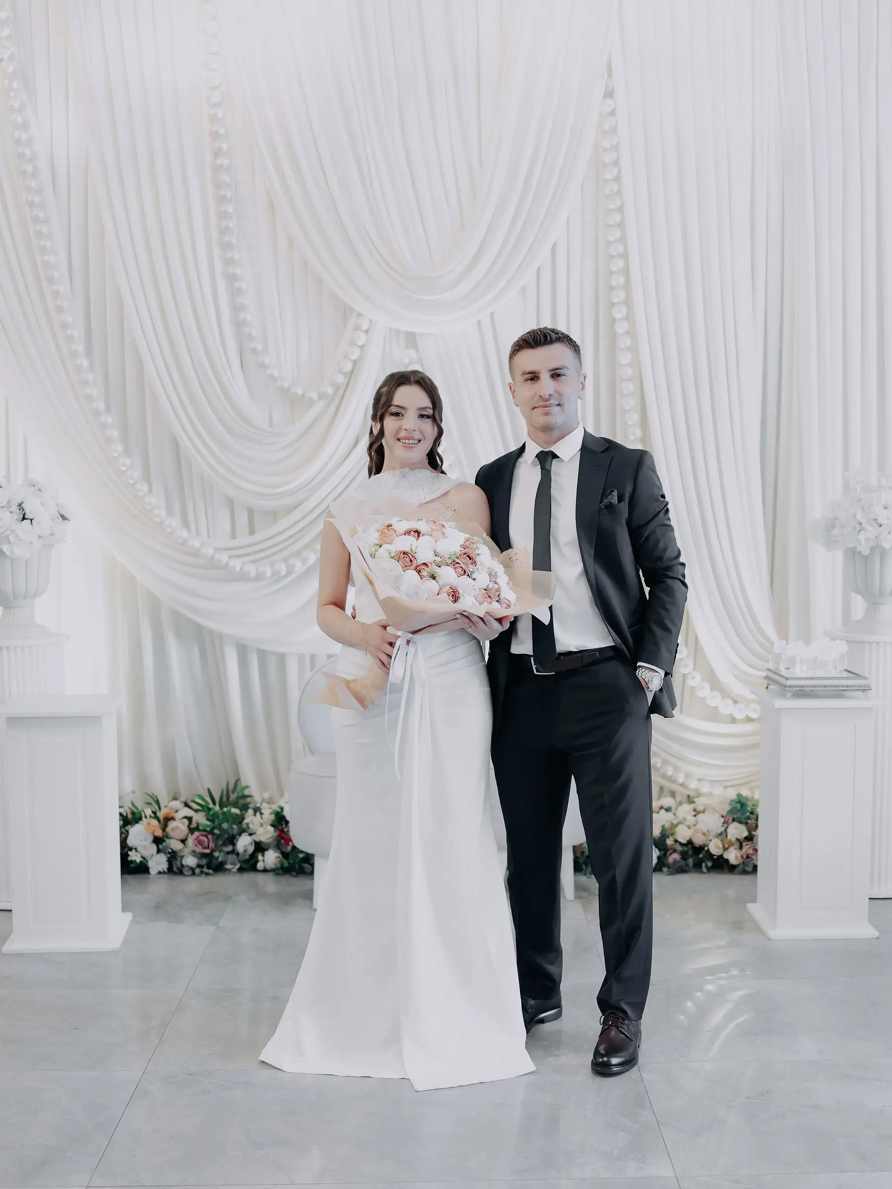 Gelin ve damat, beyaz drapeli fonun önünde yumuşak renkli buketle poz veriyor