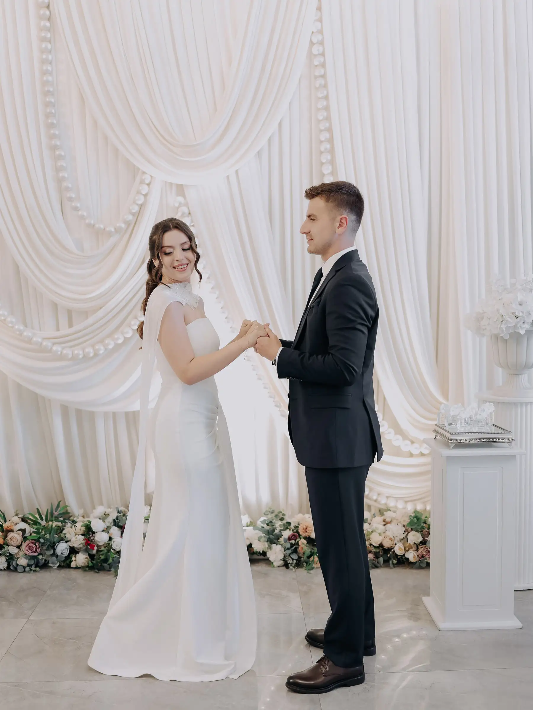 Gelin ve damat, beyaz drapeli fonun önünde karşılıklı el ele duruyor