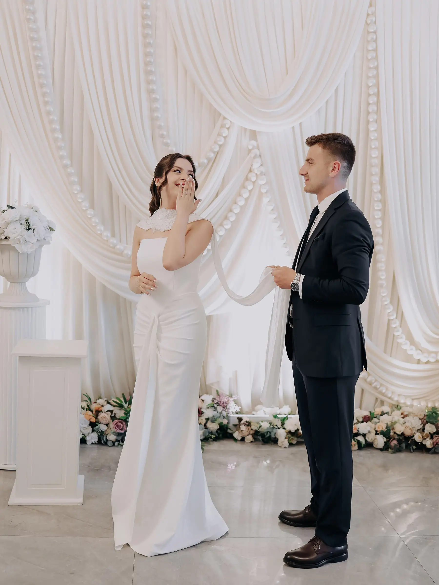 Gelin gülerek elini yüzüne götürüyor, damat karşısında duruyor; arkada beyaz drapeli fon