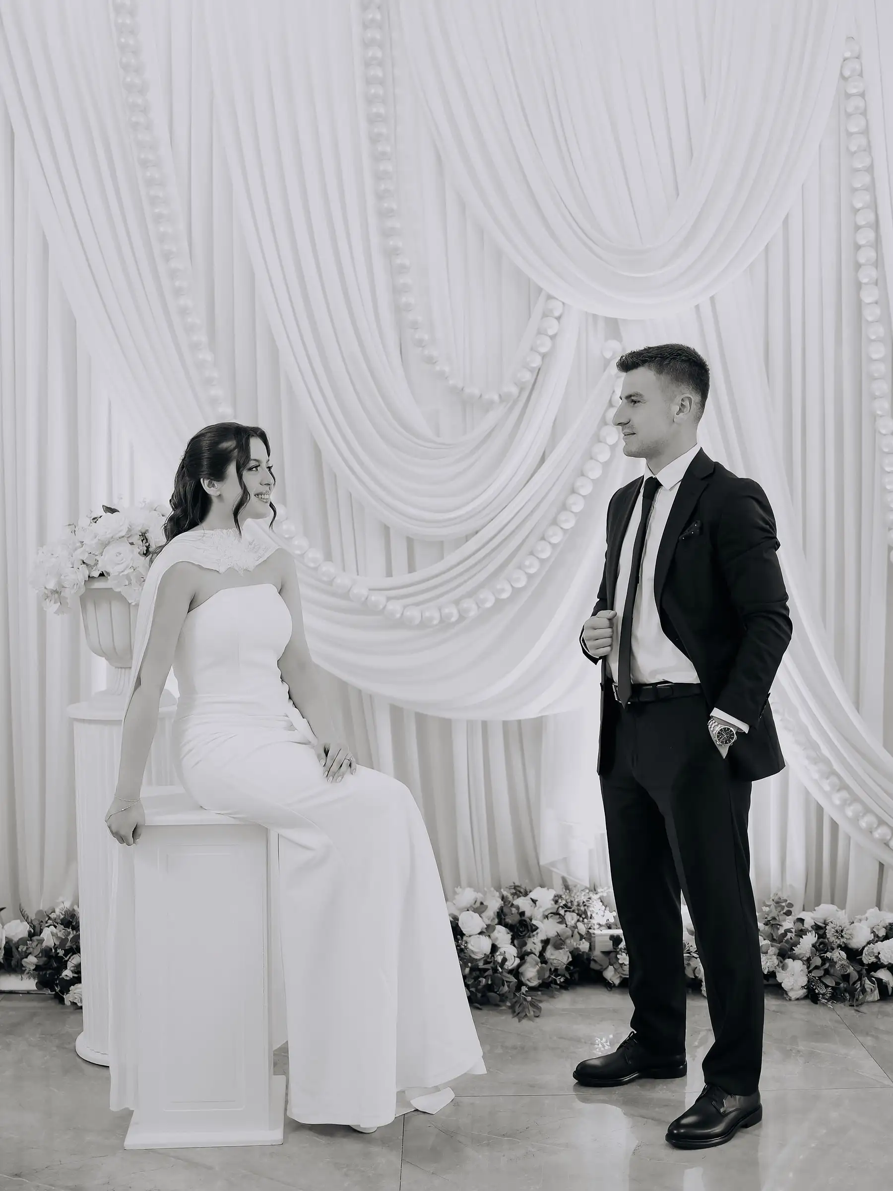 Gelin beyaz bir kaidede oturuyor, damat yanında ayakta; arkada beyaz drapeli fon ve çiçek şeridi