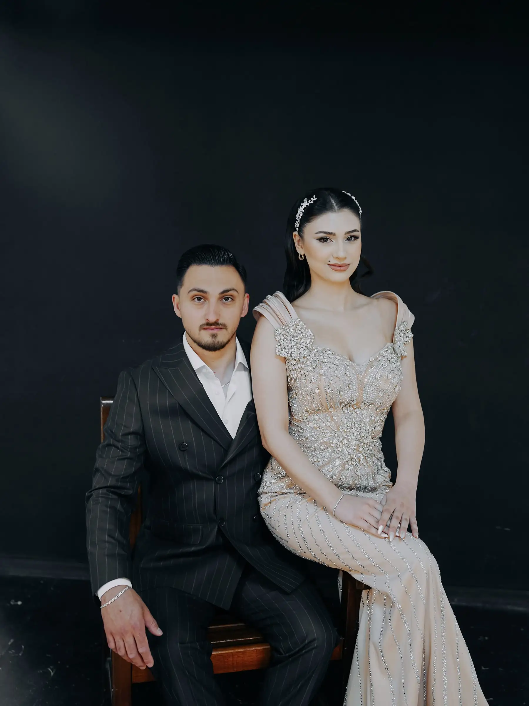 Gelin ve damat, koyu fonun önünde yan yana oturuyor; gelinin taş işlemeli elbisesi, damadın çizgili takımı