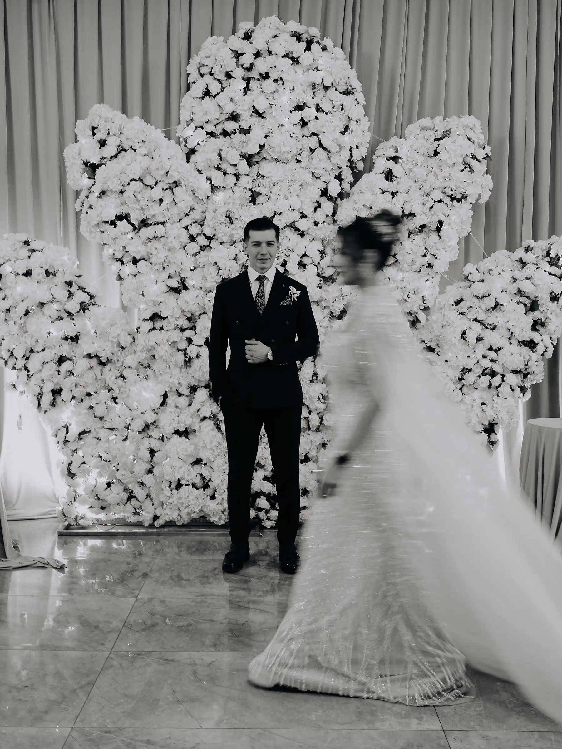 Siyah beyaz karede gelin uzun pelerinle yürüyor, damat karşıdan kameraya bakıyor