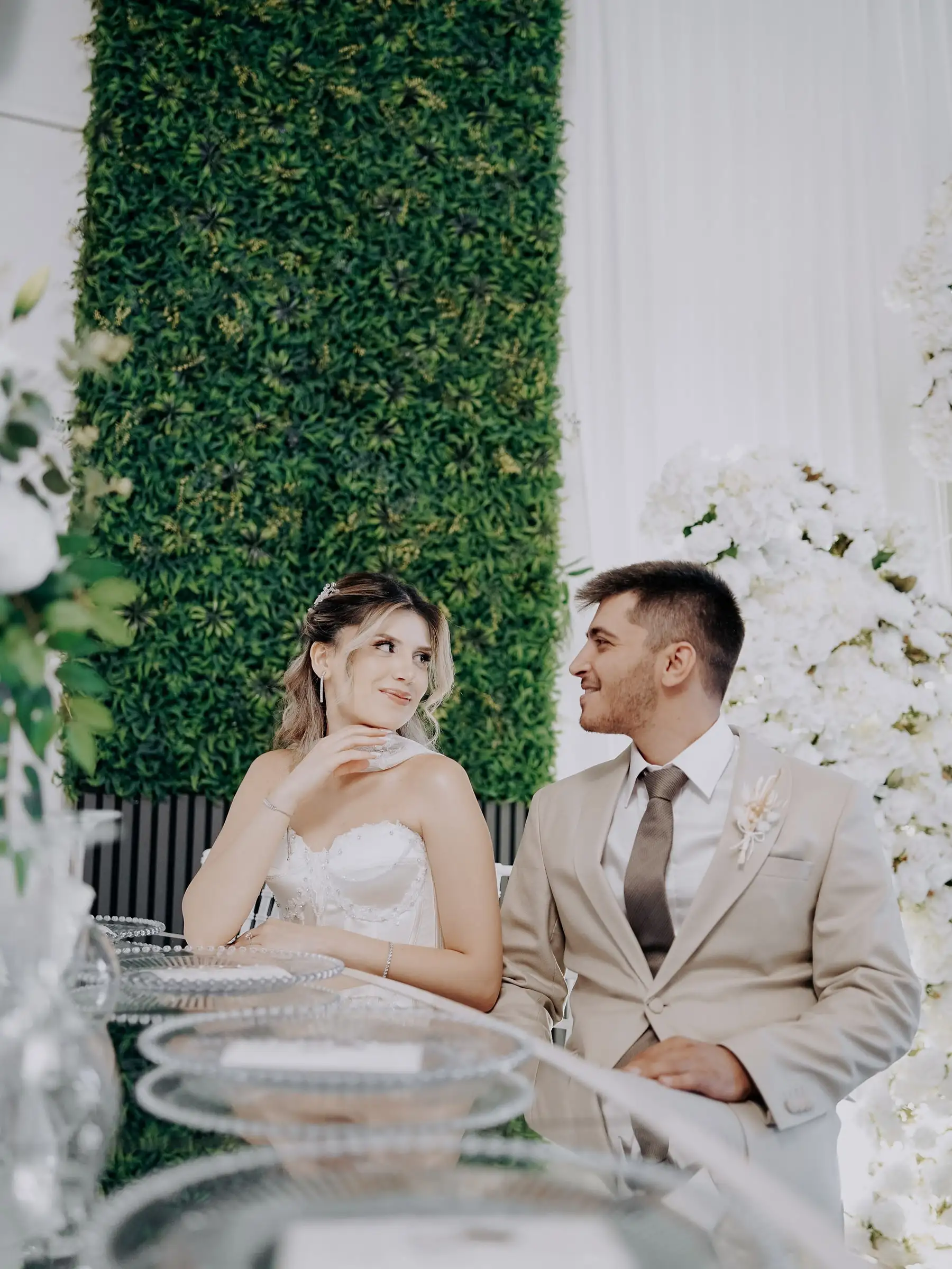 Gelin ve damat, yeşil duvarın önündeki kristal tabaklı masada birbirine bakıyor