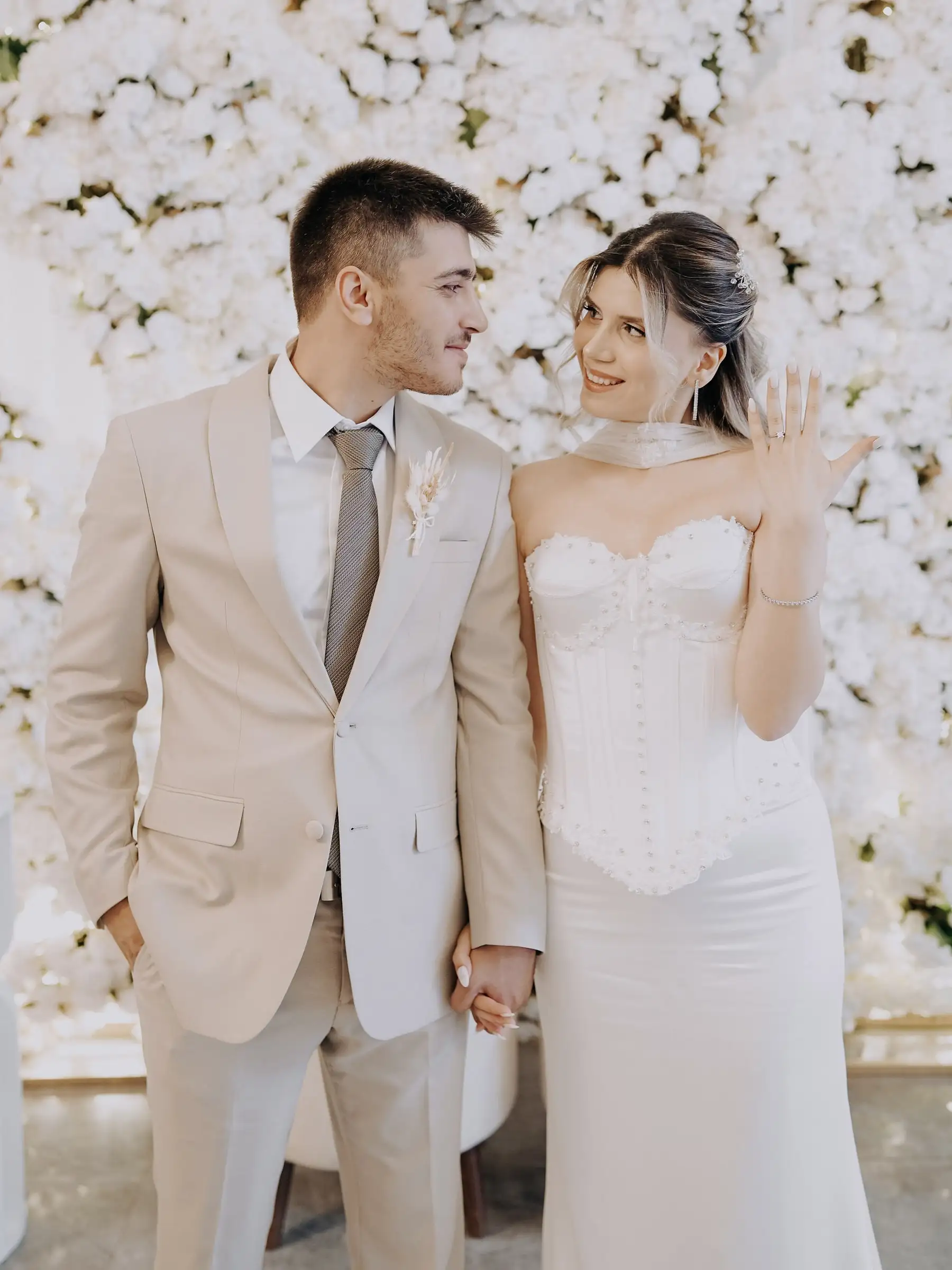 Gelin yüzüğünü gösteriyor, damat yanında; arkalarında beyaz çiçek duvarı